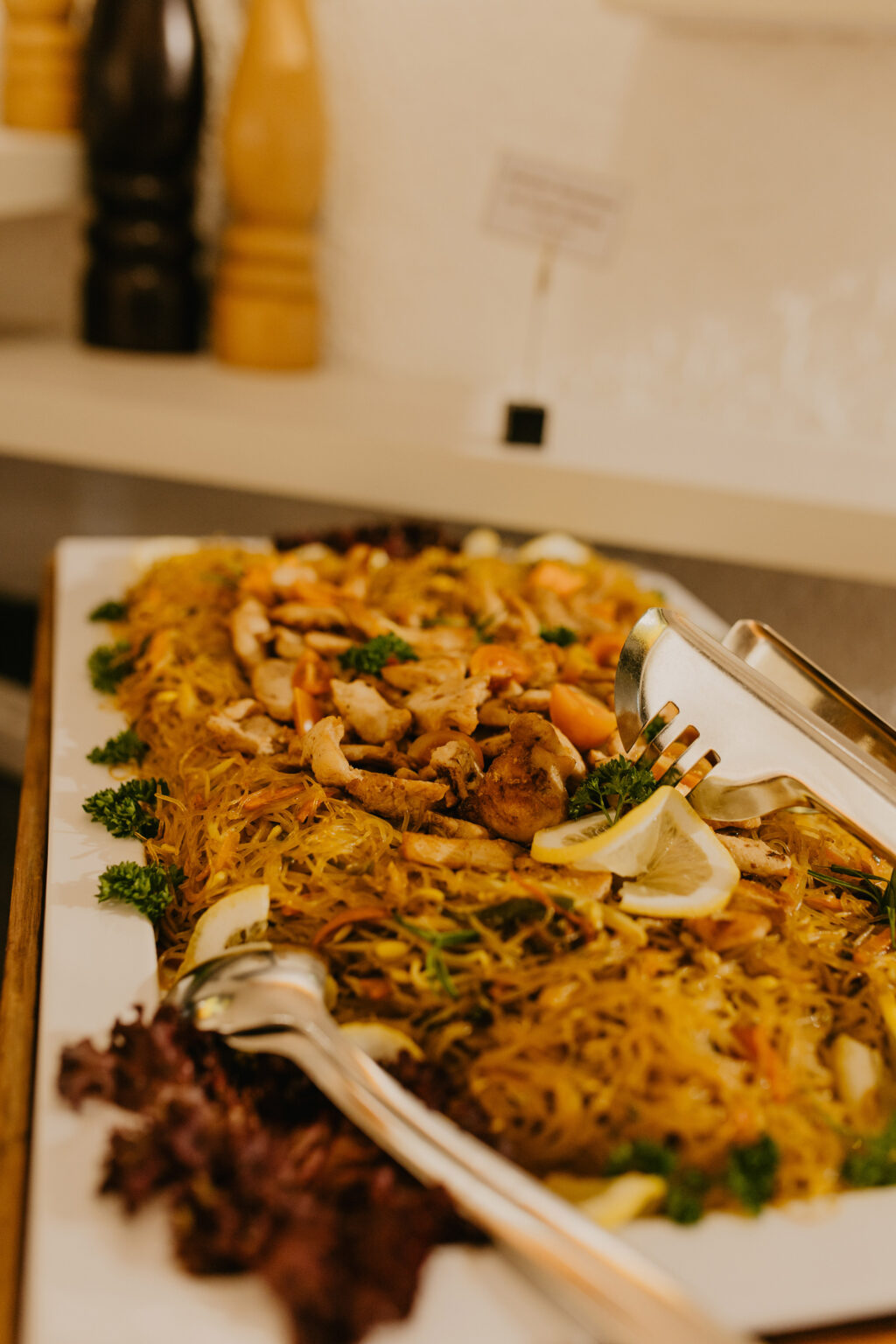Ein reichhaltiges Buffet mit Nudeln, Hähnchen, Zitronenscheiben und Servierzangen im Bereich Essen und Trinken.