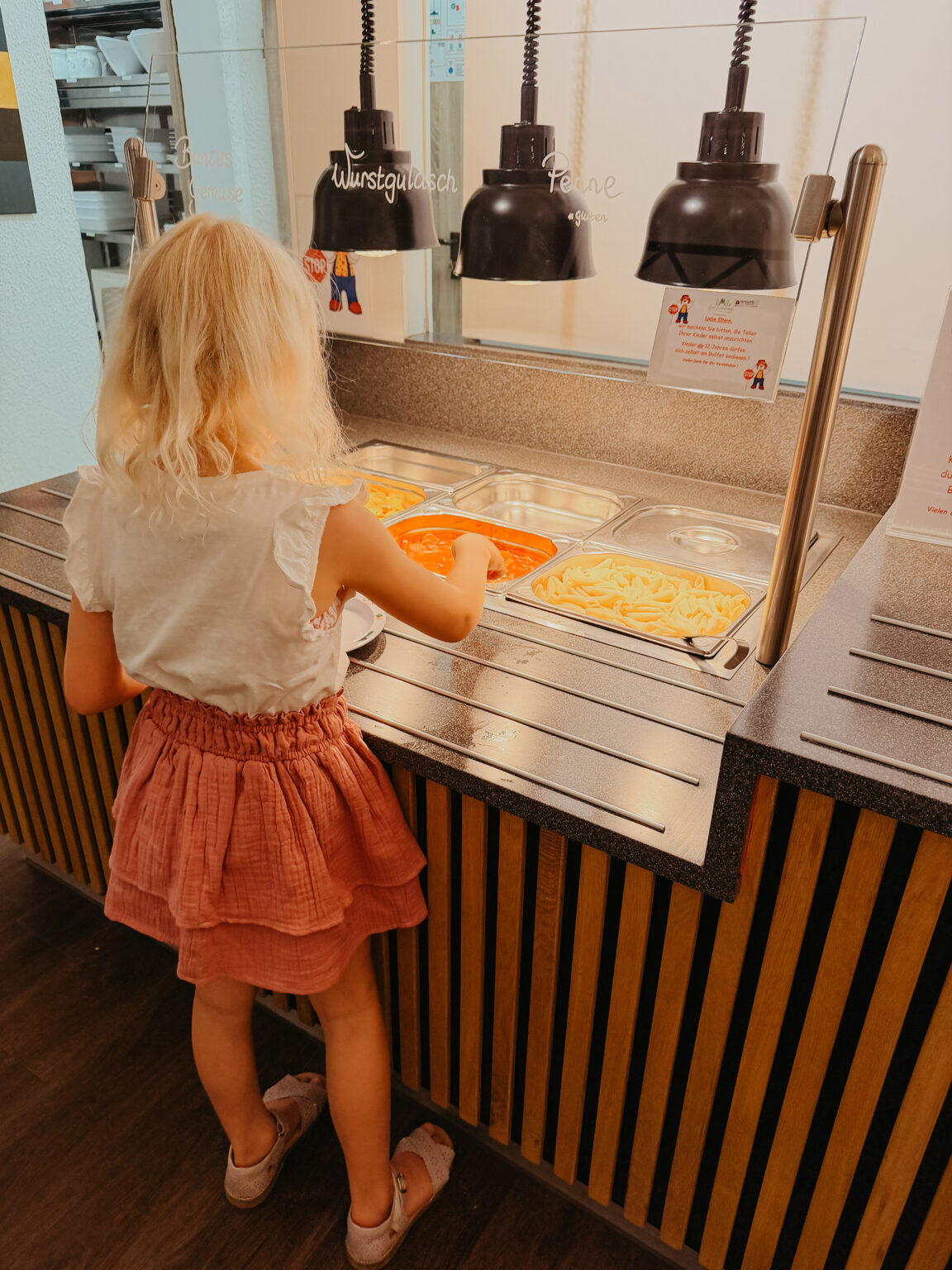 Ein blondes Mädchen im rosa Rock nimmt am Kinderbuffet bei Gut Landegge Essen aus den Warmhaltebehältern.