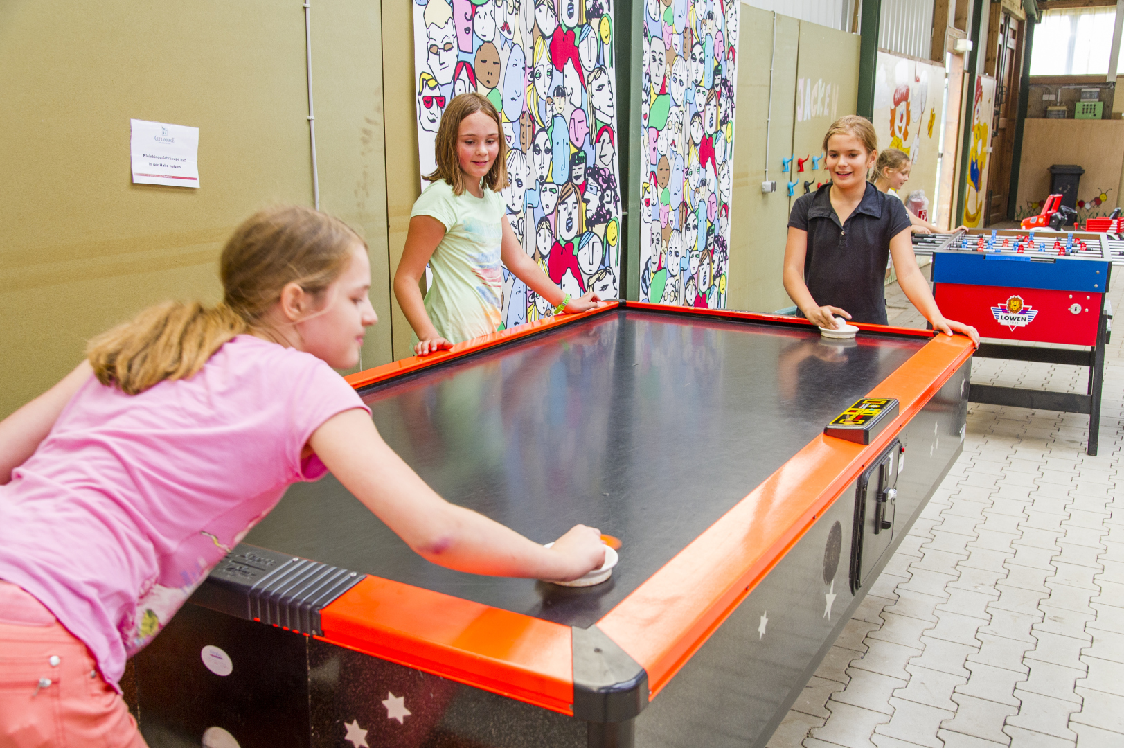 Drei Mädchen spielen begeistert Air Hockey an einem bunten Tisch in einer hellen Halle mit farbigen Wandbildern und einem Kickertisch daneben im Raum.