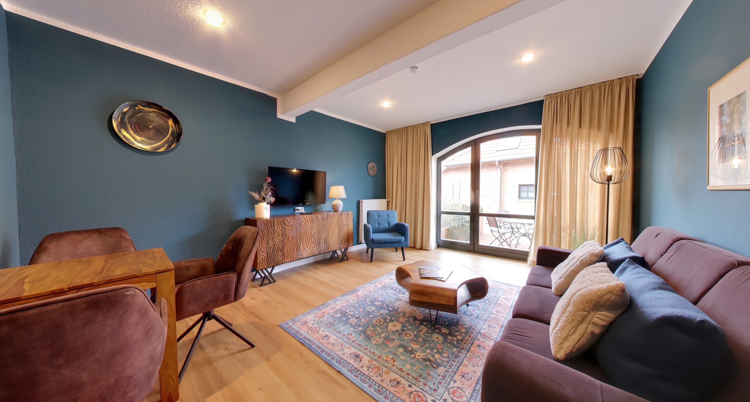 Ein gemütlich eingerichtetes Wohnzimmer mit blauen Wänden, Sofa, Sessel, TV, gemustertem Teppich und großem Rundbogenfenster zum Innenhof.
