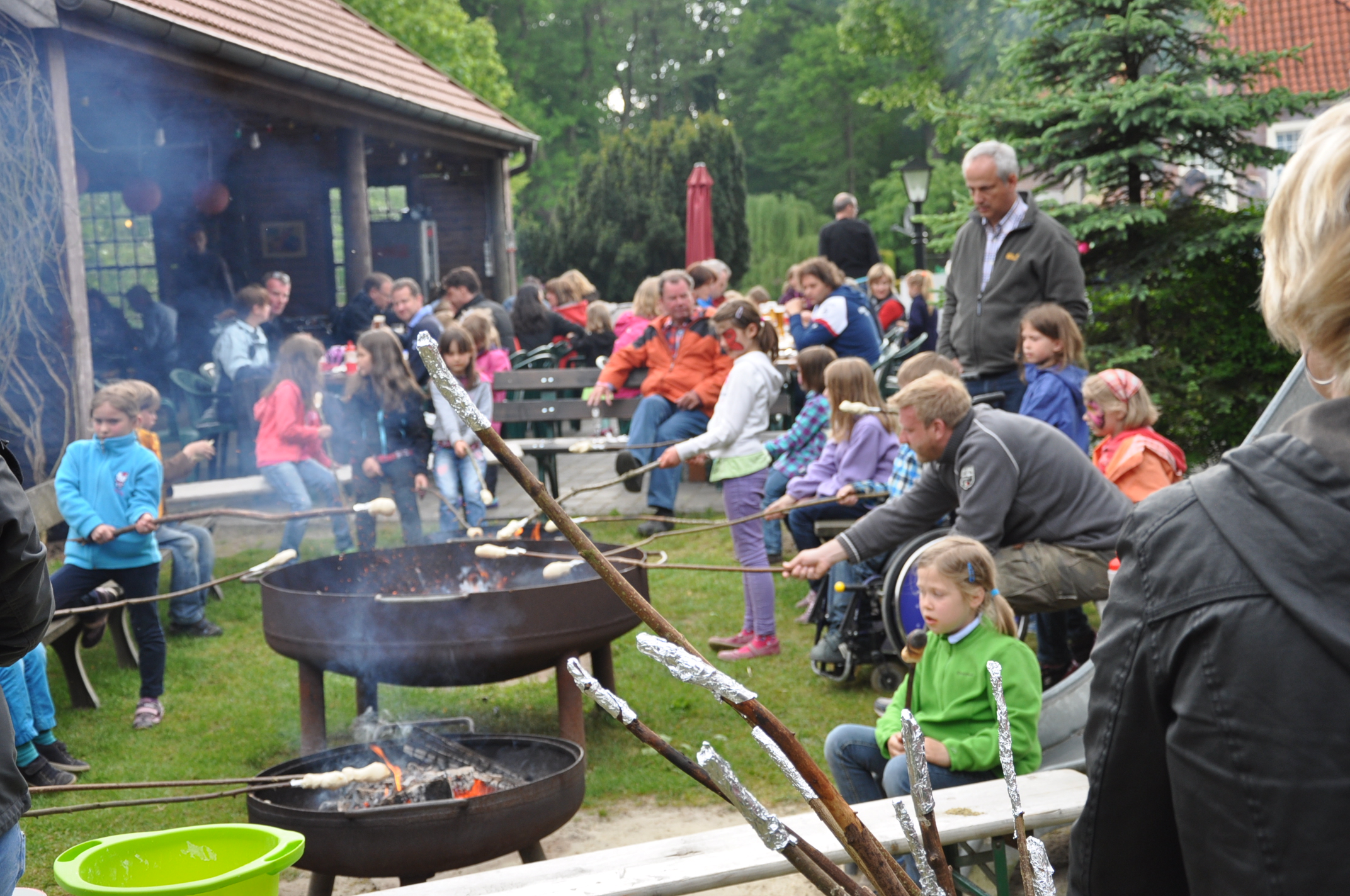 Im Familotel rösten Kinder und Erwachsene gemeinsam Stockbrot über offenen Feuerstellen auf der Wiese neben Sitzbänken und Bäumen im Garten.
