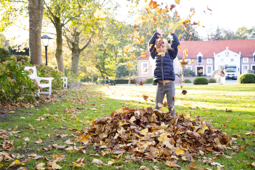 Ein blondes Kind wirft im herbstlichen Garten mit dem Arm hoch ein Blattdekor in die Luft, während hinter ihm ein Gutshof mit rotem Dach im Licht steht.