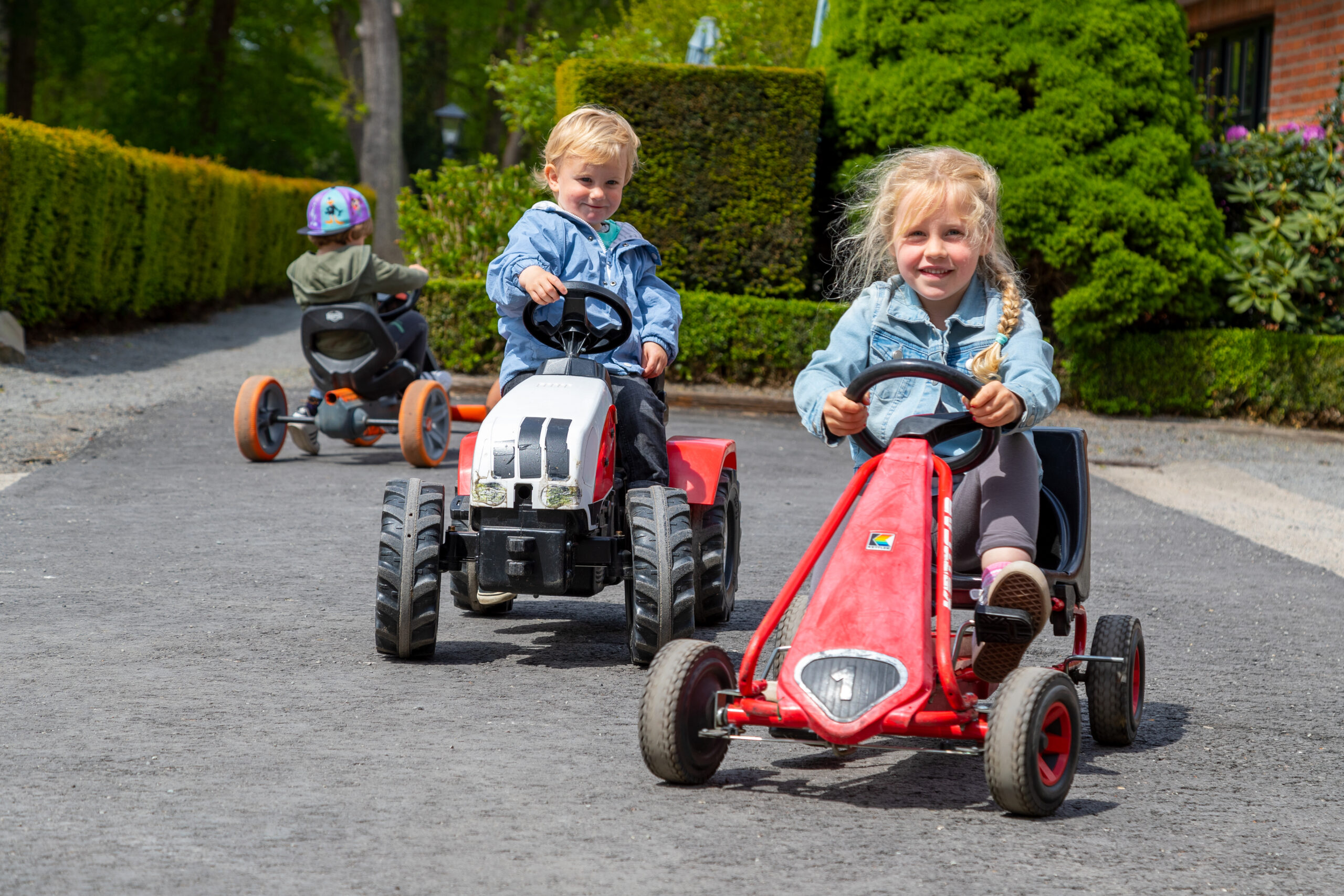 Auf einem grünen Weg fahren drei Kinder mit Tretkarts, vorn ein blondes Mädchen im Jeanshemd, dahinter zwei weitere auf dem Hof von Gut Landegge.