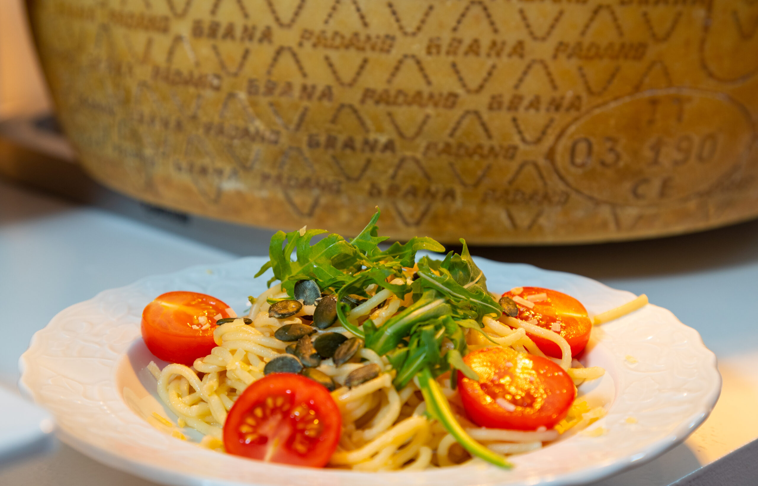 Spaghetti-Salat mit Kirschtomaten, Rucola und Kürbiskernen liegt vor einem großen Parmesanlaib auf einem weißen Teller im warmen Küchenlicht bereit.