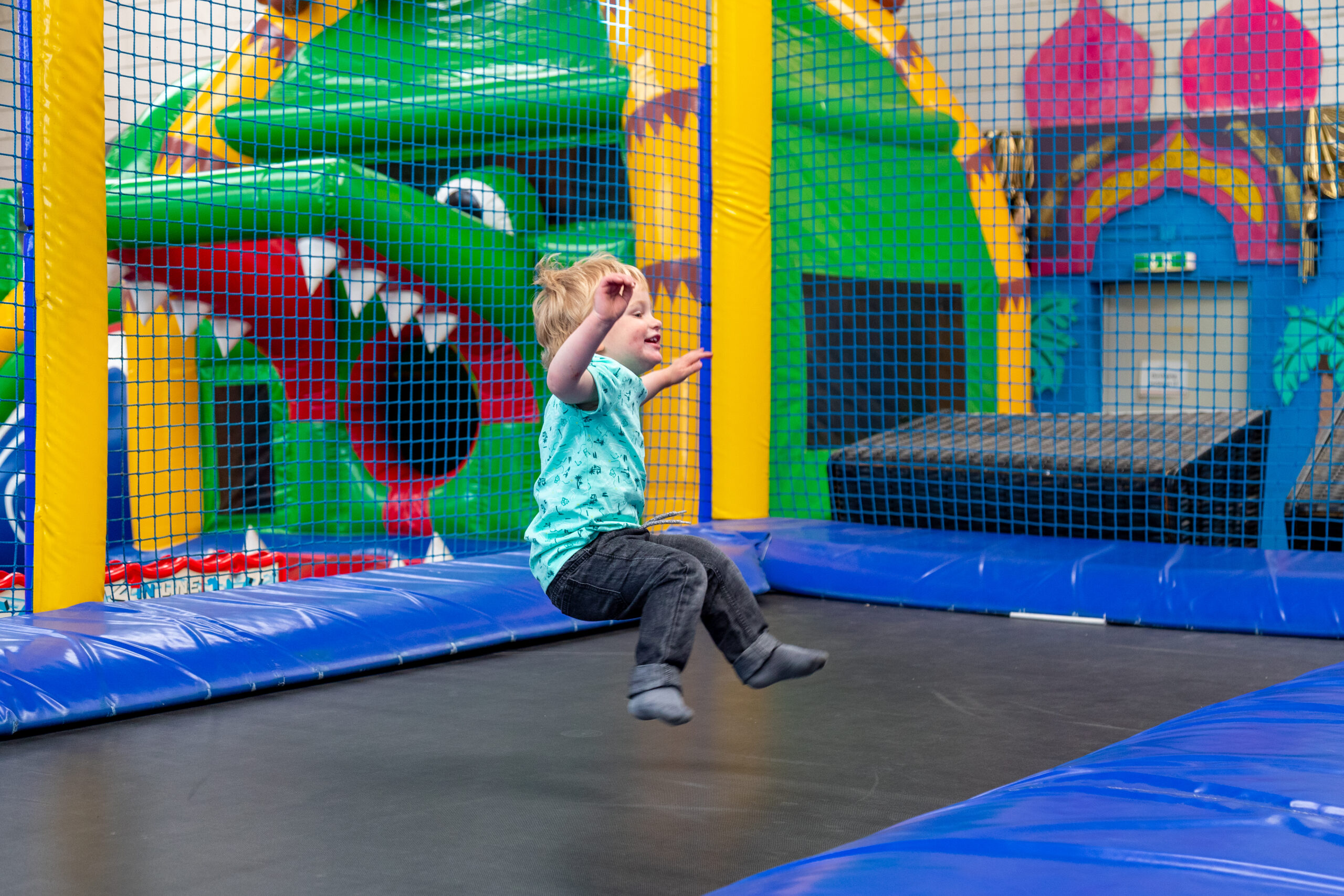 Ein blondes Kind springt lachend auf einem Trampolin zwischen bunten Netzen und Polstern in einer farbenfrohen Indoor-Spielanlage bei Gut Landegge.