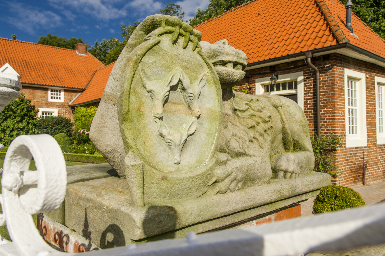 Eine steinerne Skulptur mit drei Fuchsköpfen steht vor rot gedeckten Backsteingebäuden und einem hellen Gartenzaun im sonnigen Hof.