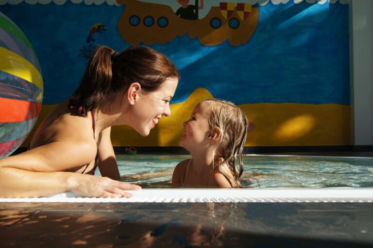 Familienhotel Gut Landegge im Emsland: kleines Mädchen lacht mit Mama im Babybecken, Hintergrund ein Wandbild mit Boot und ein bunter Wasserball