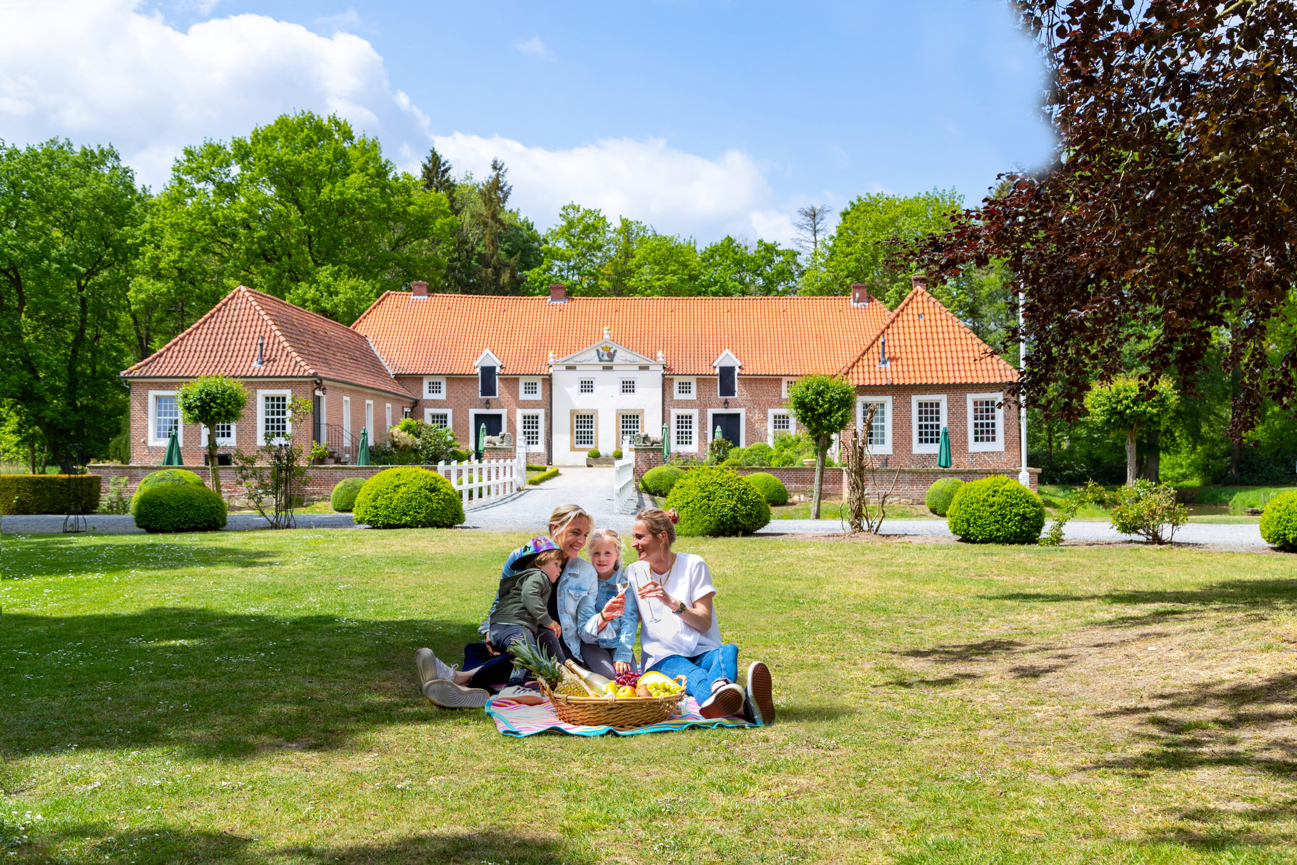 Vor dem Gut Landegge sitzt eine Familie auf einer Picknickdecke im Park vor dem Herrenhaus mit rotem Ziegeldach bei Sonnenschein und Obstkorb.