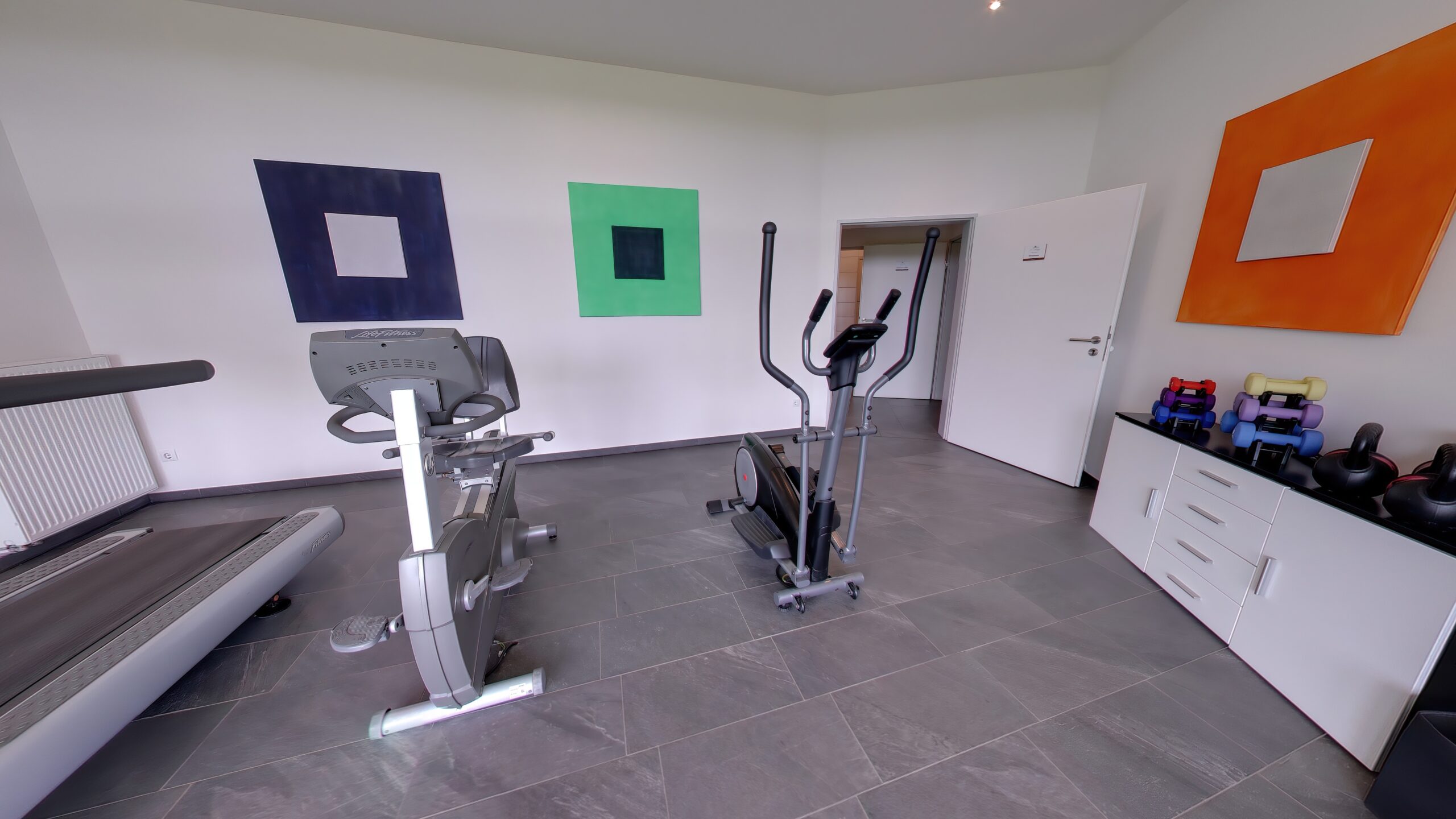 Moderner Fitnessraum mit Laufband, Crosstrainer und Hantelbank, ergänzt durch bunte Wandbilder, Gewichte und eine offene Tür zum Nebenraum.