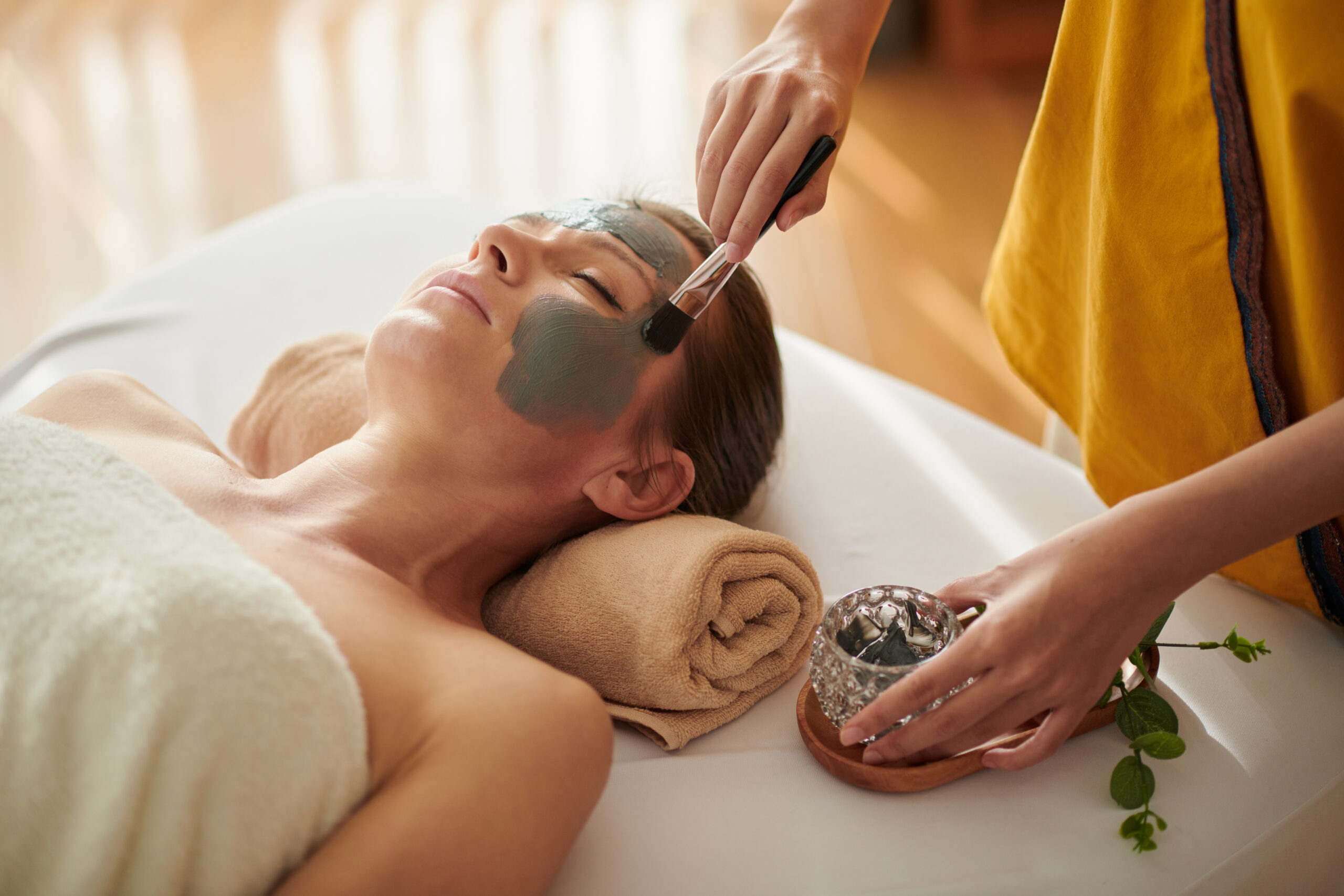 Eine Frau erhält in der Kosmetikbehandlung eine grüne Gesichtsmaske, während eine Hand mit dem Pinsel das Gesicht im Spa sanft bestreicht.
