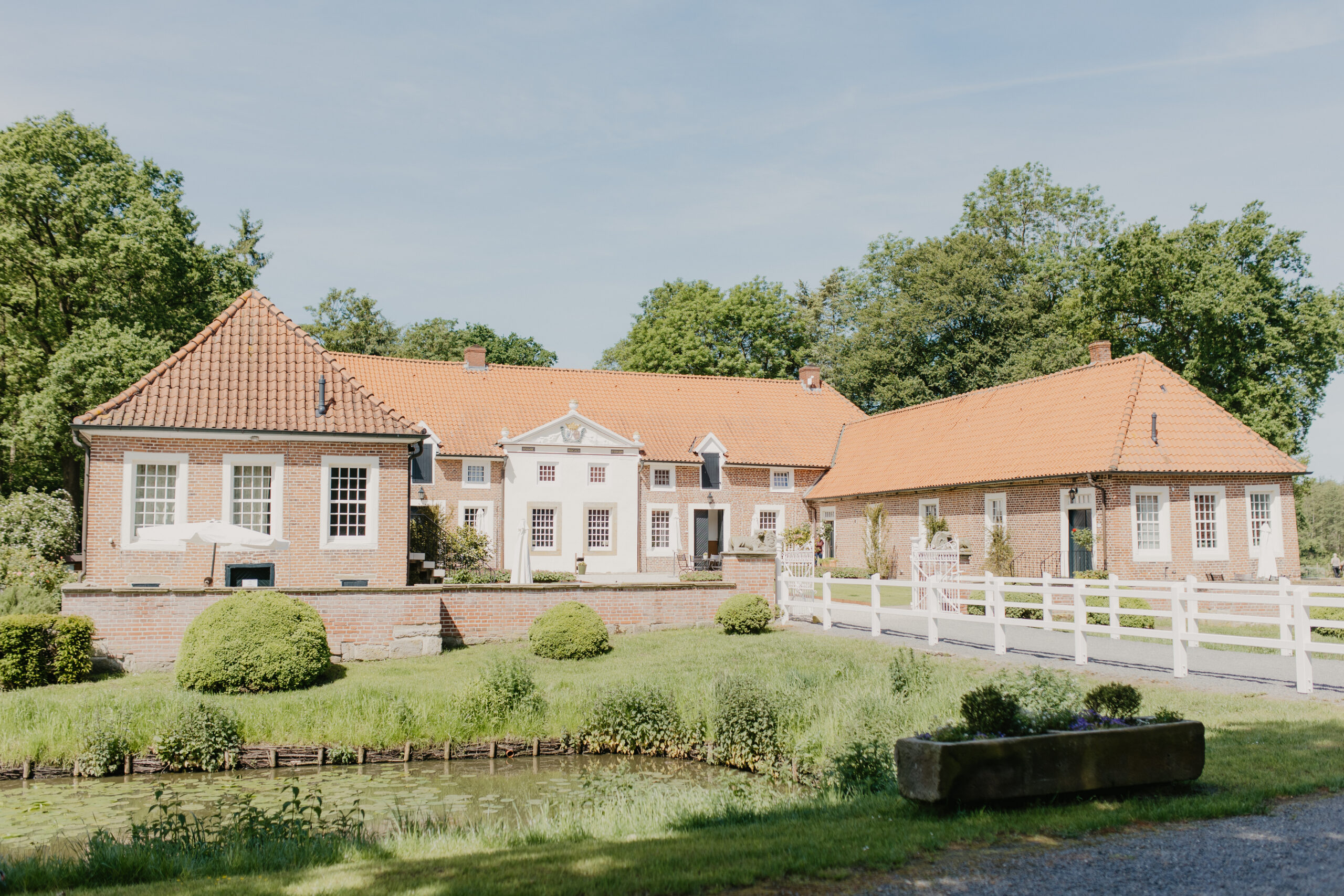 Die historische Backsteinanlage mit roten Ziegeldächern, weißem Zaun und Teich liegt ruhig im Grünen in Landegge.
